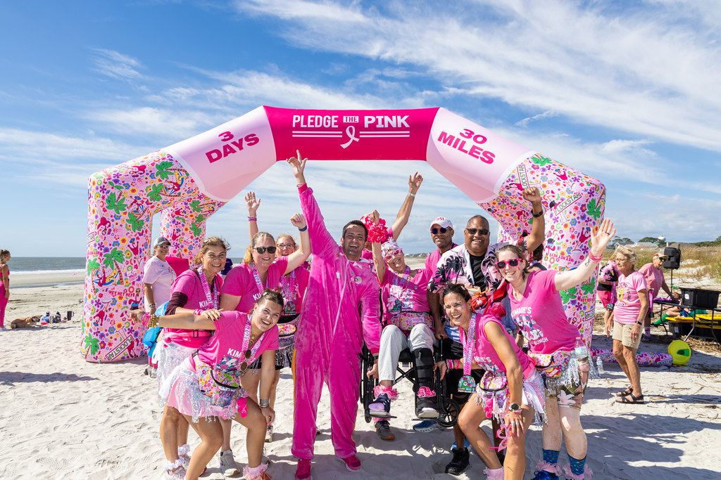 A team in pink cheering on the beach under the inflatable Pledge the Pink "3 Days, 30 Miles" arch