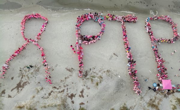 Aerial view of hundreds of people in pink forming a pink ribbon and the letters PTP on a beach