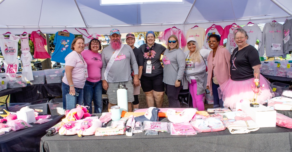 Nine smiling volunteers in pink behind the merchandise table under a white tent hung with Pledge the Pink shirts, one in a pink tutu and one with a pink beard.