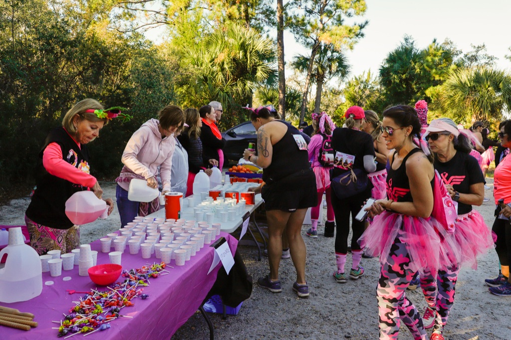 Volunteers pouring drinks into rows of cups at a purple pit-stop table while registrants in pink tutus stop by.