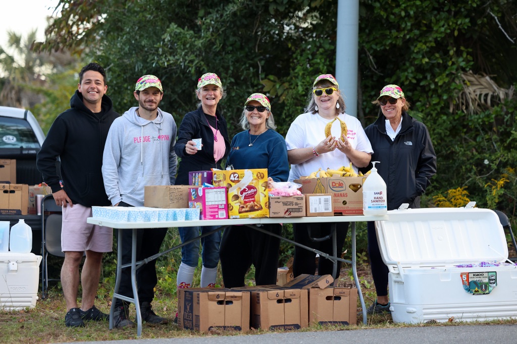 Six volunteers in matching pink camo caps behind a snack table stacked with cups, bananas and cookies, one holding up a bunch of bananas.